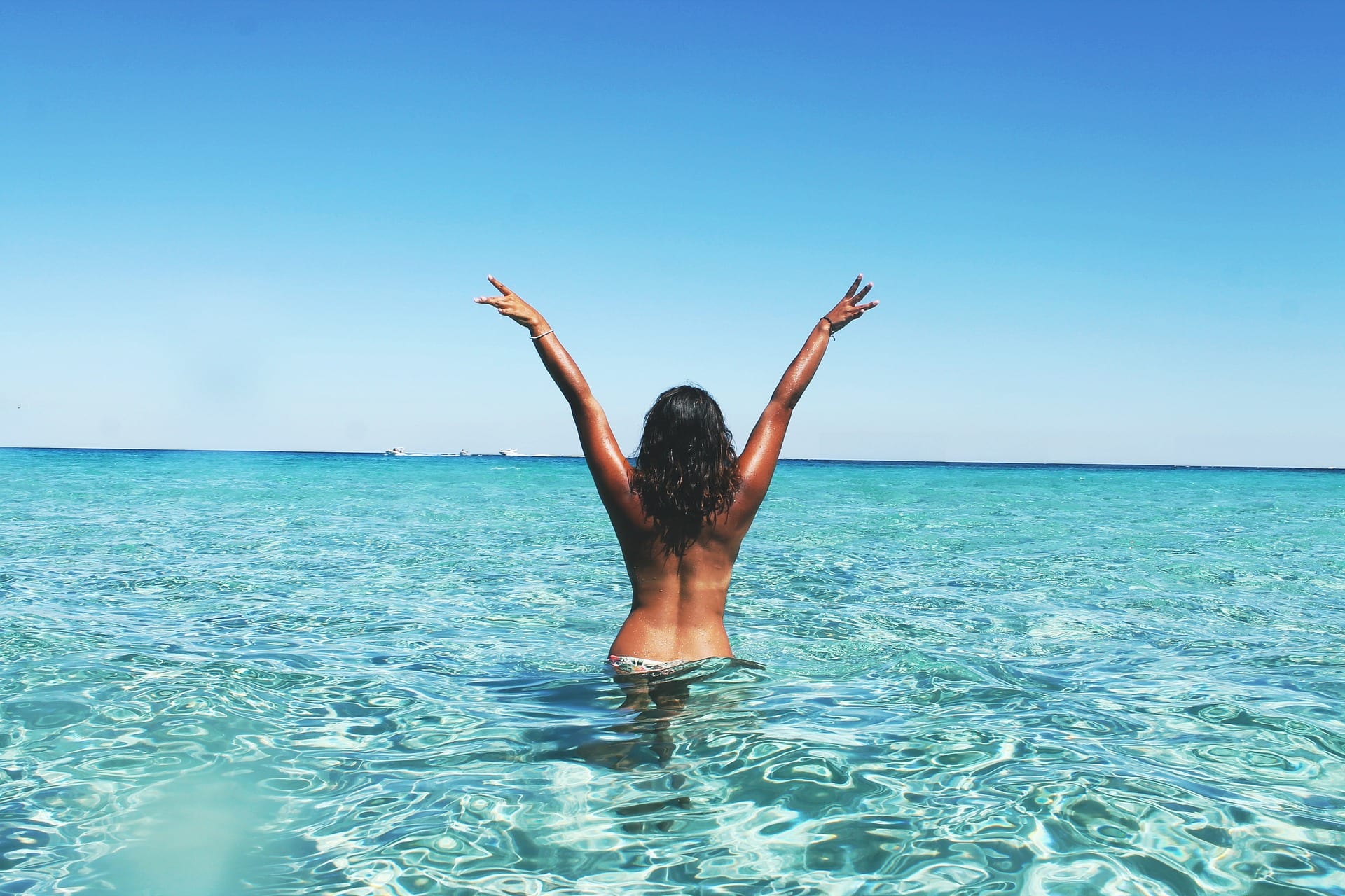 A dark-skinned woman standing hip-deep in the sea, back to the camera, arms thrown up in triumph.